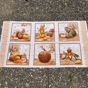K&ouml;nnte beinhalten: Stoffpaneel mit sechs Quadraten mit herbstlichen Illustrationen. Jedes Quadrat zeigt K&uuml;rbisse und Waldtiere wie Eichh&ouml;rnchen, Waschb&auml;ren und F&uuml;chse. Die Farbpalette umfasst warme Braun- und Oranget&ouml;ne, die die Jahreszeit widerspiegeln.