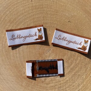 Puede incluir: Tres etiquetas de tela blanca con bordes marrones y texto. Dos etiquetas presentan la palabra "Lieblingsstück" en escritura marrón y una ilustración de zorro. La tercera etiqueta tiene un diseño marrón. Todas las etiquetas están sobre una superficie de madera.