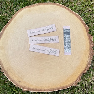 Puede incluir: Tres pequeñas etiquetas rectangulares de papel marrón con el texto "handgemachte Glück" impreso en gris. Las etiquetas están sobre una superficie de madera.