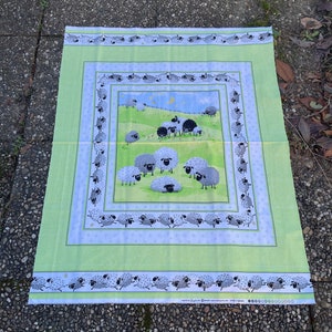 May include: A green fabric panel with a white border and a repeating pattern of black sheep. The center of the panel features a larger image of a field with black sheep and a crescent moon.