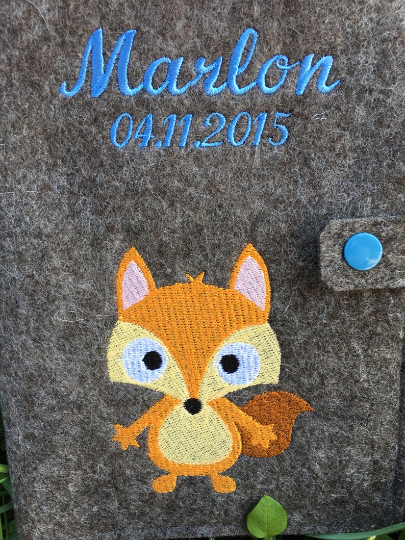Puede incluir: Una bolsa de fieltro gris con cierre de bot&oacute;n azul. La bolsa tiene un zorro bordado con pelaje naranja, vientre amarillo y ojos azules. El texto "Marlon 04.11.2015" est&aacute; bordado en azul en la bolsa.