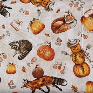 Peut inclure: Un tissu crème avec un motif répété de citrouilles, de renards, de ratons laveurs et d'un épouvantail fait de citrouilles. Les citrouilles sont orange et jaune, avec des accents bruns. Les renards sont orange et marron, et les ratons laveurs sont gris et noirs.