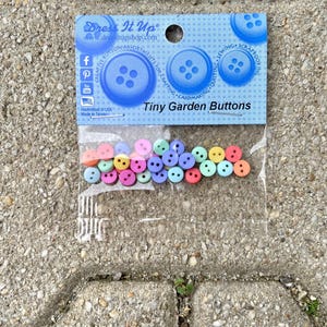 Könnte beinhalten: Eine Packung mit kleinen bunten Gartenknöpfen. Die Verpackung ist blau und weiß mit den Worten "Dress It Up" und "Tiny Garden Buttons".