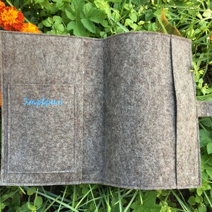 Puede incluir: Un portapasaporte de fieltro gris con cierre de bot&oacute;n azul y un bolsillo en el interior con el texto "Simplypass" bordado en azul.