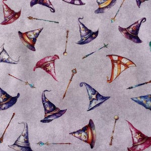Peut inclure: Un fond violet avec un motif répété de chapeaux de sorcier et de baguettes magiques colorés. Les chapeaux sont de styles et de couleurs variés, notamment violet, bleu, rouge et vert. Les baguettes sont argentées et ont une étoile à l'extrémité.