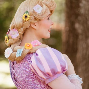 Könnte beinhalten: Eine Frau mit langen blonden Haaren, die zu einem Zopf geflochten sind und mit bunten Blumen-Haarspangen verziert sind. Sie trägt ein lavendelfarbenes Kleid mit rosa gestreiften Ärmeln. Der Hintergrund ist verschwommen.