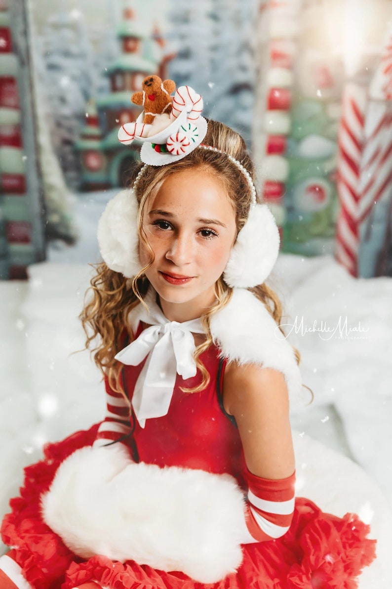 May include: A young girl wearing a red dress with white fur trim and white earmuffs. She is wearing a festive headband with a gingerbread man, candy canes, and a cup of hot chocolate. The background is a snowy winter scene.