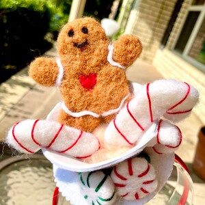 May include: A gingerbread man with a red heart on his chest sits in a white cup with candy canes and peppermint candies. The cup is attached to a red headband.