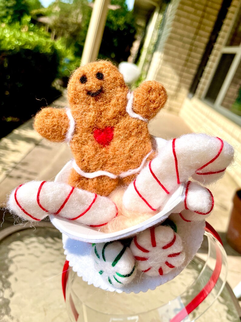 May include: A gingerbread man with a red heart on his chest sits in a white cup with candy canes and peppermint candies. The cup is attached to a red headband.