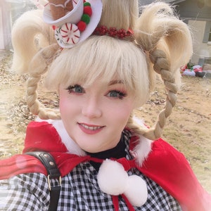 May include: A woman wearing a red cape and a black and white gingham dress with a white pom-pom trim. Her hair is styled in two braids with a gingerbread man and candy cane hat.