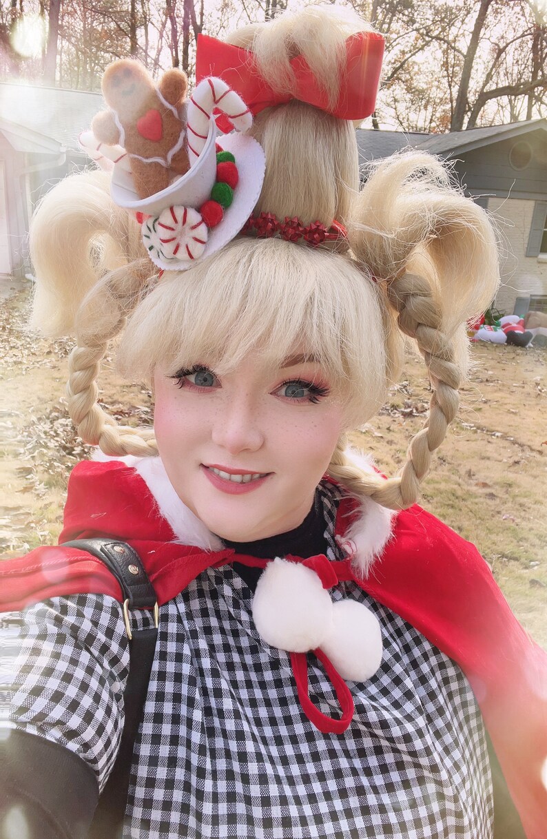 May include: A woman wearing a red cape and a black and white gingham dress with a white pom-pom trim. Her hair is styled in two braids with a gingerbread man and candy cane hat.