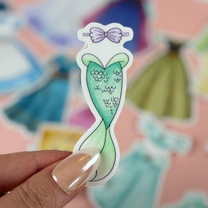 May include: A mermaid tail sticker with a green scale design and a light green tail. The sticker has a purple bow at the top and is held by a person. Other stickers are in the background.