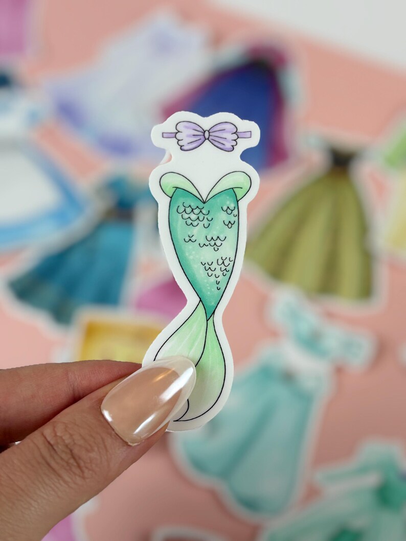 May include: A mermaid tail sticker with a green scale design and a light green tail. The sticker has a purple bow at the top and is held by a person. Other stickers are in the background.