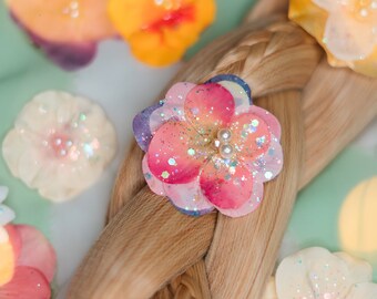 May include: A braid of light brown hair with several small, glittery flower hair clips. The clips are in various colors, including pink, yellow, and white.