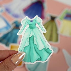 May include: A sticker featuring a teal-colored dress with a seashell necklace. The dress has long sleeves and a flowing skirt. Other stickers of dresses are in the background. The sticker is held by a person.