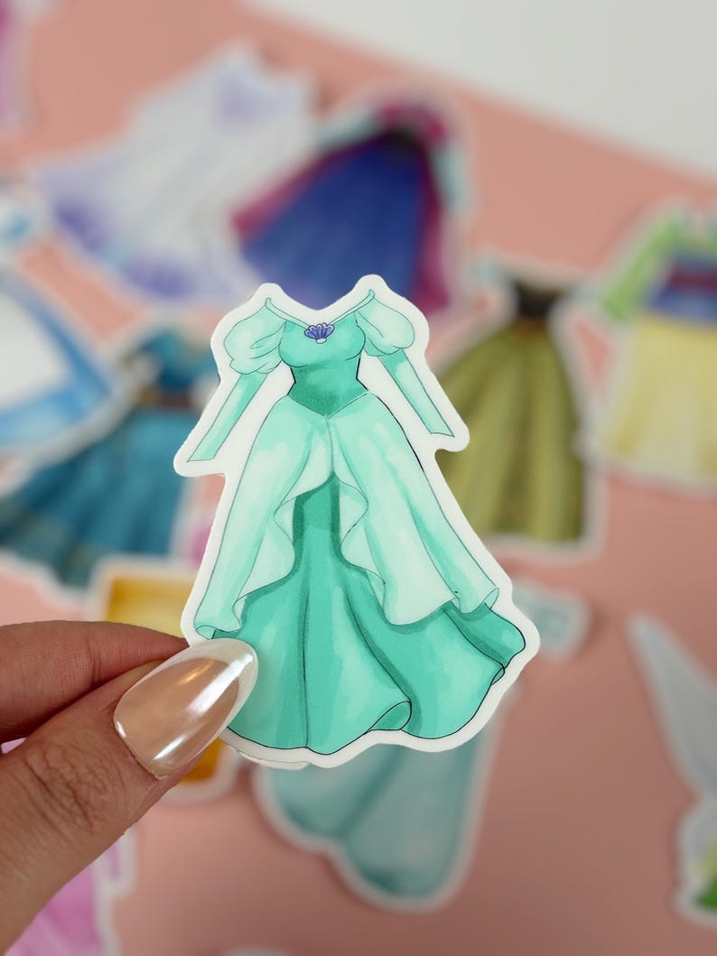 May include: A sticker featuring a teal-colored dress with a seashell necklace. The dress has long sleeves and a flowing skirt. Other stickers of dresses are in the background. The sticker is held by a person.
