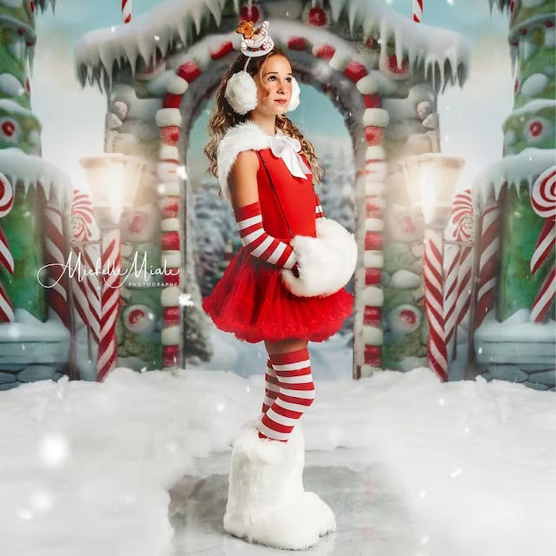 May include: A young girl wearing a red and white striped dress, white fur boots, and a white fur headband with a red bow. She is standing in front of a candy cane themed backdrop.