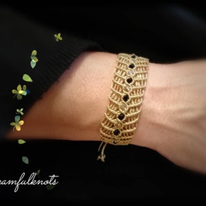 Könnte beinhalten: Ein goldfarbenes Makramee-Armband mit schwarzen und klaren Perlen. Das Armband hat ein gewebtes Design. Das Armband befindet sich an einem Handgelenk. Der Text "Dreamfulknots" befindet sich in der unteren linken Ecke.