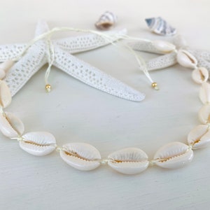 Könnte beinhalten: Eine weiße Muschelkette mit einer weißen Schnur und goldenen Perlen. Die Kette besteht aus Kaurischnecken und ist auf eine weiße Schnur gefädelt.