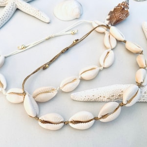 Könnte beinhalten: Eine weiße Muschelkette mit braunem Band und goldenen Perlen. Die Kette besteht aus Kaurischnecken und ist auf einer natürlichen Schnur aufgefädelt.