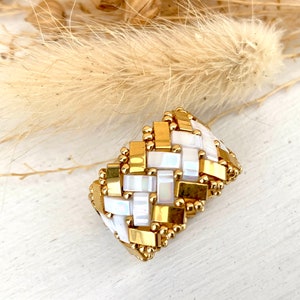 Op de afbeelding: Een gouden en witte kralenring. De ring heeft een geweven ontwerp met rechthoekige gouden en witte kralen. Deze ring is een statement-stuk, perfect om een vleugje elegantie toe te voegen. De ring is ongeveer 2.5 cm breed.