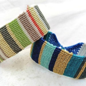 May include: Two macrame bracelets with colourful striped patterns. One bracelet features green, grey, and gold stripes. The other bracelet has blue, teal, and gold stripes. The bracelets are handmade and sit on a white surface.