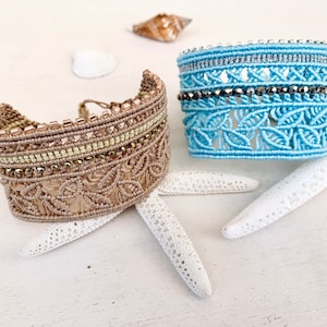 Peut inclure: Deux bracelets en macramé. L'un est beige avec des perles dorées et vertes, l'autre est bleu clair avec des perles argentées et gris foncé. Les deux sont de style manchette large. Les bracelets sont exposés sur une surface blanche avec des étoiles de mer et des coquillages.