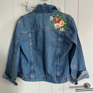 May include: A blue denim jacket featuring a floral embroidery on the back. The design showcases red, pink, yellow, and white flowers with green foliage. The jacket has a standard collar, button fastenings, and rolled-up sleeves.