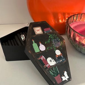 Peut inclure: Une boîte à bijoux noire en forme de cercueil avec un couvercle ouvert révélant des bagues. La boîte est décorée d'un motif d'étagère peint avec un crâne, des livres, des plantes et des bouteilles. Un grand vase orange et une bougie rose sont en arrière-plan.