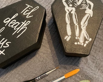 May include: Two small black wooden boxes shaped like coffins. One box has the words "Til death do us part" written in white paint. The other box has a white painted image of two skeletons holding each other and a red heart above them. Two paintbrushes are also in the image.