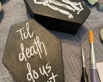 May include: Two black wooden coffin-shaped boxes. One box features a white skeleton couple embracing with a red heart above them. The other box has the words "Til death do us part" written in white.