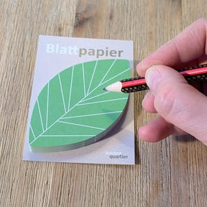 Könnte beinhalten: Ein grüner, blattförmiger Notizblock mit dem Text "Blattpapier" und "aussen. quartier" ist abgebildet. Eine Hand hält einen Bleistift mit roten und schwarzen Akzenten und zeigt auf den Notizblock. Der Notizblock liegt auf einer Holzoberfläche.