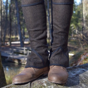 May include: Brown tweed leg warmers with a dark blue trim, worn over brown leather shoes and blue jeans. The leg warmers extend to the knees, offering warmth and style. The background shows a natural outdoor setting.