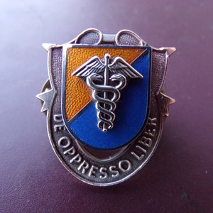 May include: A silver-toned metal shield-shaped pin with a medical caduceus emblem. The shield has orange and blue sections, with the words "DE OPPRESSO LIBER" in a circular pattern around the edge.