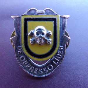May include: A silver pin with a yellow and black shield featuring a skull and crossbones. The shield is surrounded by a silver band with the text "DE OPPRESSO LIBER".