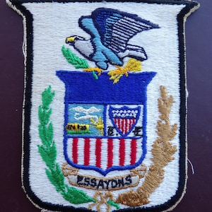 May include: A white embroidered patch with a blue eagle perched on a shield. The shield has a blue background with a smaller shield inside, featuring a white star and stripes. The larger shield has red and white stripes and a green wreath around it. The text "Essayons" is embroidered on a white ribbon.
