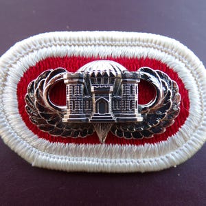 May include: An embroidered patch with a red oval center, bordered by white stitching. A silver emblem featuring a castle with wings is centered on the patch. The patch is likely a military insignia or emblem.