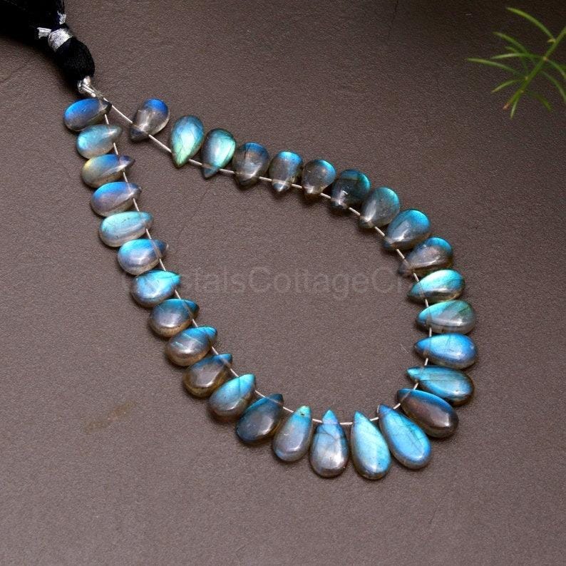 May include: A strand of teardrop-shaped labradorite beads. The beads display a range of blues, grays, and iridescent flashes. The beads are strung on a thin, silver-colored wire, with a black tassel at the top. The beads are approximately 1 cm in length.