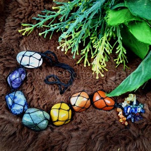 May include: A seven-stone chakra necklace with clear, purple, blue, green, yellow, orange, and red stones. Each stone is wrapped in black string and connected to a black cord.