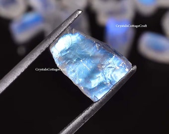 May include: A close-up of a blue iridescent gemstone held by tweezers. The gemstone is clear and has a rainbow sheen. CrystalsCottageCraft is written on the image.