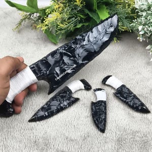 Może przedstawiać: Kolekcja obsydianowych noży i grot&oacute;w strzał. Duży n&oacute;ż ma białą owiniętą rękojeść i napis "CrystalsCottageCraft". Mniejsze groty strzał mają białe owinięte rękojeści. Obsydian ma błyszczący, czarny wygląd.