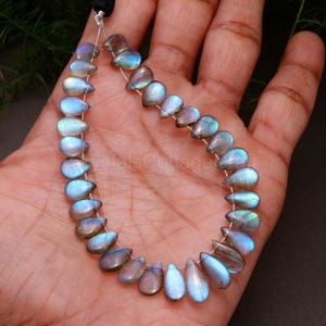 May include: A strand of teardrop-shaped labradorite beads. The beads display a mix of blue, gray, and iridescent colors. The beads are strung together on a thin, clear wire. The beads are approximately 0.5 inches in length.