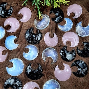 May include: A collection of crescent moon shaped stones in various colours, including black, pink, and blue. The stones are arranged on a brown furry surface.