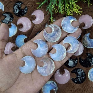 Può includere: Una collezione di pietre di opale bianco iridescente a forma di mezzaluna. Le pietre sono di varie dimensioni e hanno un colore bianco latteo con iridescenza blu e viola.