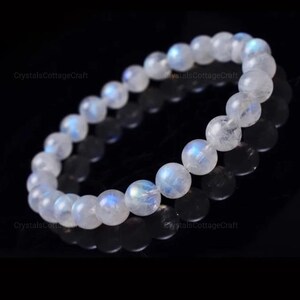 May include: A white moonstone bracelet with iridescent blue and white tones. The bracelet is made of round beads and is strung on an elastic cord.