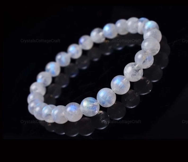 May include: A white moonstone bracelet with iridescent blue and white tones. The bracelet is made of round beads and is strung on an elastic cord.