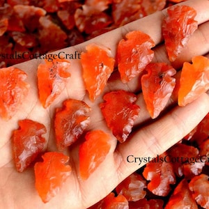 以下が含まれることがあります： オレンジのカーネリアンの矢じりのコレクション。一部は赤みがかった色合い。手の中にあります。矢じりは様々なサイズと形をしています。画像にはCrystalsCottageCraftと書いてあります。