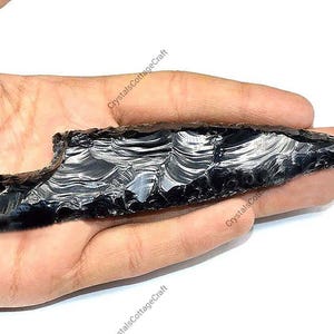 Può includere: Una punta di freccia in ossidiana nera lucidata, con una superficie lucida e una punta affilata. La punta di freccia è tenuta in una mano, evidenziando la sua consistenza e forma. Il prodotto è di CrystalsCottageCraft.