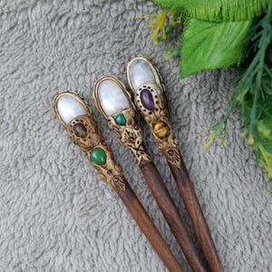 May include: Three wooden hair sticks with decorative tops featuring iridescent white stones and colorful gemstones. The sticks are brown and have intricate carvings.