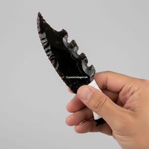 May include: A black obsidian knife with a serrated edge and a white wrapped handle. The blade is a deep, glossy black, and the handle is wrapped with white string. The knife is held in a hand against a neutral background.